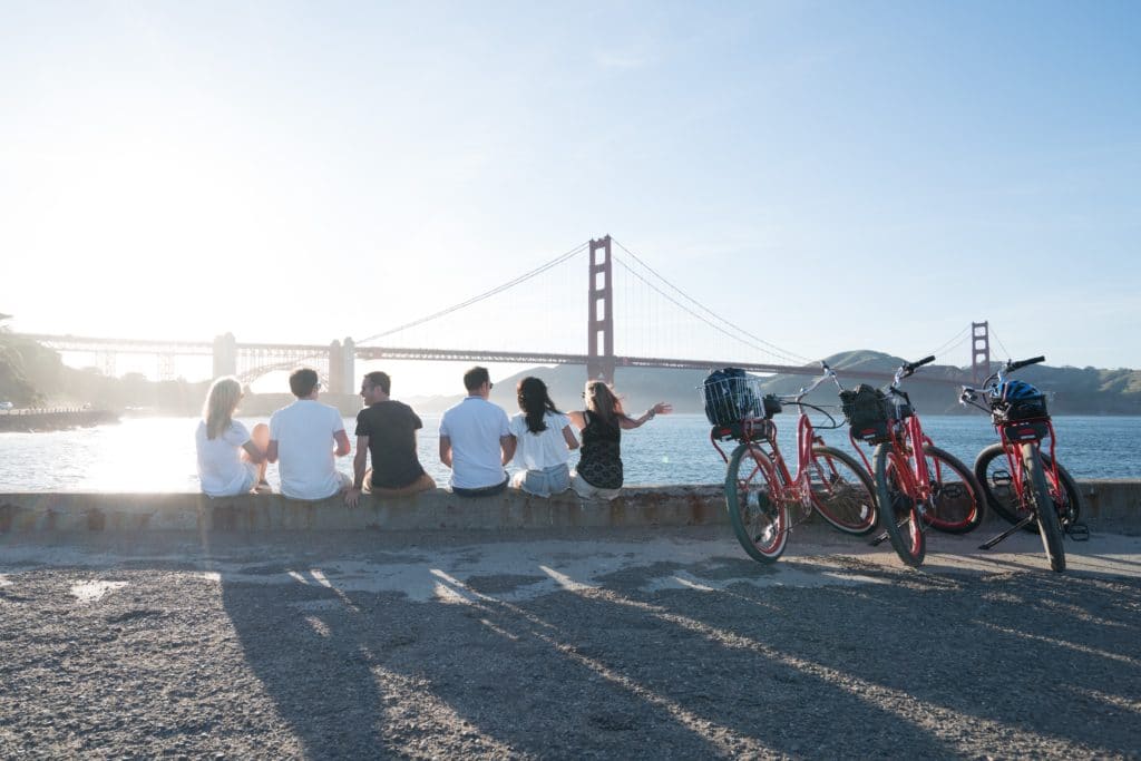 6 friends sitting on a pier in view of the Golden Gate Bridge, beside three bikes.