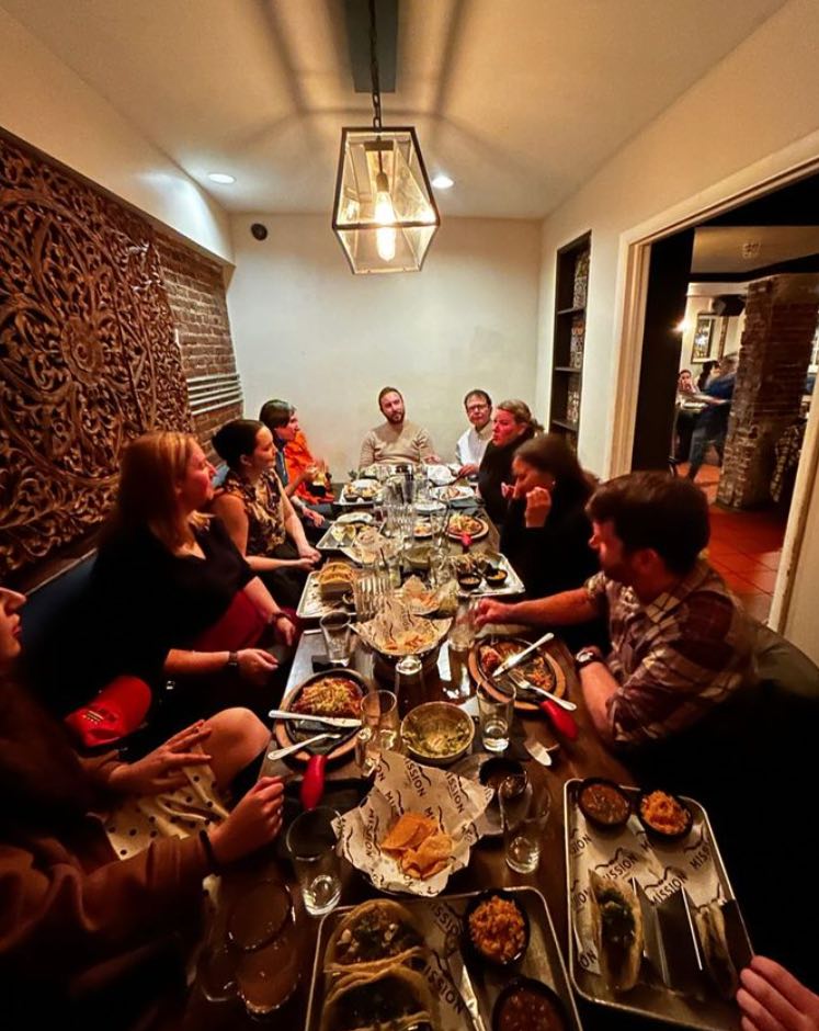 A group of 9 people gathered around a table eating and talking